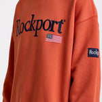 Burnt orange cotton crew neck sweatshirt with embroidered Rockport logo and american flag at chest, worn by model